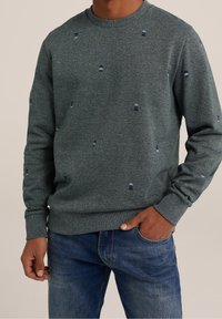 Man som bär en mörkgrå tröja med rund hals och små mönstrade prickar, i kombination med blå jeans, med en hand i fickan och står mot en enfärgad bakgrund.