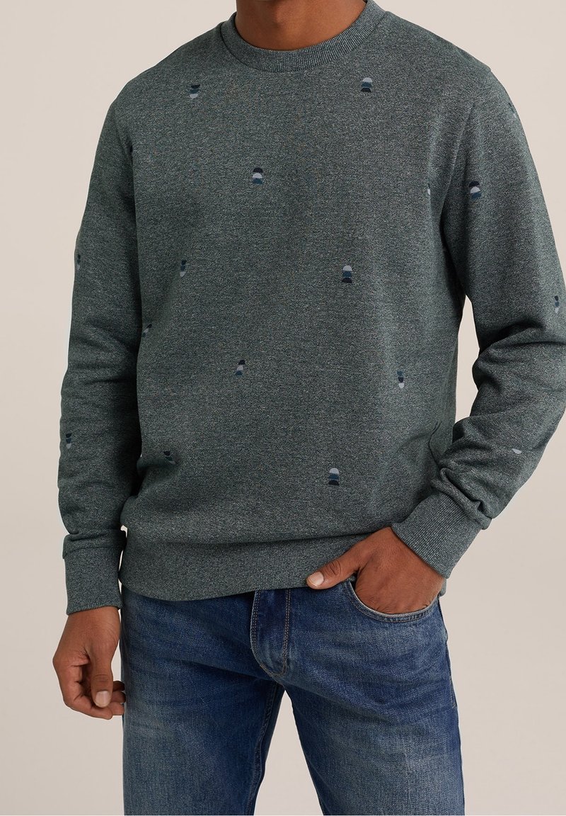 Man som bär en mörkgrå tröja med rund hals och små mönstrade prickar, i kombination med blå jeans, med en hand i fickan och står mot en enfärgad bakgrund.