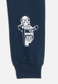 Dunkelblaue Jogginghose mit elastischen Bündchen, die ein weißes Druckdesign eines LEGO-Ninja-Charakters in dynamischer Pose zeigt.