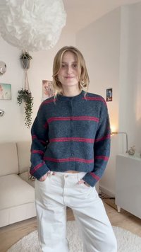 Mėlynas megztinis su raudonomis juostelėmis, apvaliu iškirpimu ir mygtukų uždarymu. Sukombinuotas su baltais plačiais kelnais, kurie pabrėžia laisvą dydį.
