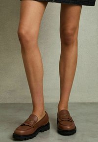 Brune læder loafers med frynset detalje og guldfarvet accent på vampen, sat på en tykk sort gummisål. Synlige ben iført en nederdel.