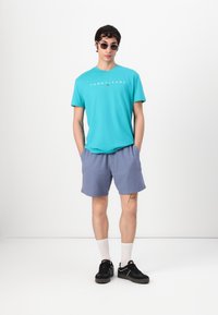 Tyrkysové tričko s logom "TOMMY JEANS", svetlomodré šortky, čierne tenisky a biele ponožky do polovice lýtok. Minimalistický dizajn na jednoduchom pozadí.