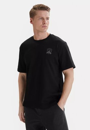 Uomo dalla pelle chiara e capelli corti che indossa una maglietta nera con un piccolo logo Beverly Hills, mani nelle tasche, guarda di lato.