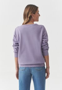 Felpa lilla con maniche lunghe e polsini a costine, vista dal retro. Abbinata a jeans blu. Tessuto morbido, design casual.