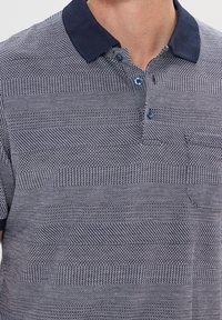 Polo shirt bleu marine avec un motif texturé et des rayures horizontales dans différentes nuances de bleu et de blanc. Fermeture à deux boutons au col.