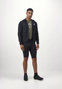 Vīrietis stāv, tērpies melnā jaku ar rāvējslēdzēju, olīvzaļā T-kreklā ar logo, melnās riteņbraucēja šortos, melnās zeķēs un melnās riteņbraukšanas kurpēs, pret baltu fona.