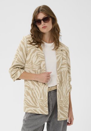 Nő bézs és fehér zebramintás blézerben, fehér inggel, szürke farmerral és sötét napszemüveggel, egy kezével a blézeren pózol.