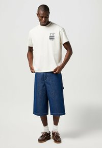 Camiseta de algodón blanca con gráfico negro de "CASEY JONES", combinada con pantalones cortos de mezclilla azules hasta la rodilla y zapatos marrones con cordones.