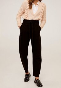 Pantalons noirs taille haute avec une taille ceinturée, associés à un cardigan à volants rose clair et une chemise blanche. Des mocassins noirs brillants complètent la tenue.