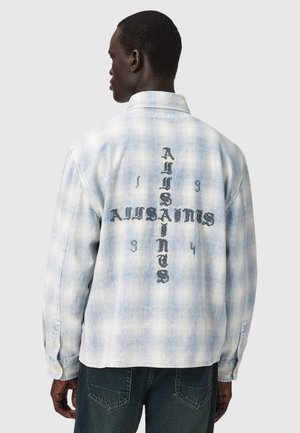 Mann trägt hellblau kariertes Hemd mit schwarzem gotischem Schriftzug "AJUSAINAS" in Form eines Kreuzes auf dem Rücken, kombiniert mit dunklen Jeans.