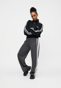 Μαύρο φούτερ με φερμουάρ και λευκές ρίγες στα μανίκια, συνδυασμένο με γκρι φόρμα με ρίγες στα πλάγια. Το μοντέλο στέκεται με σταυρωμένα χέρια.