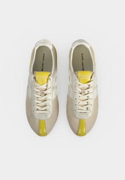 Paire de baskets crème et beige Marc Jacobs avec des accents jaunes et des lacets blancs, vue de dessus sur un fond uni.