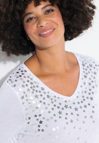 Witte V-hals t-shirt met een zilveren sterrenpatroon op de borst. Zachte stof met een relaxte pasvorm en korte mouwen.