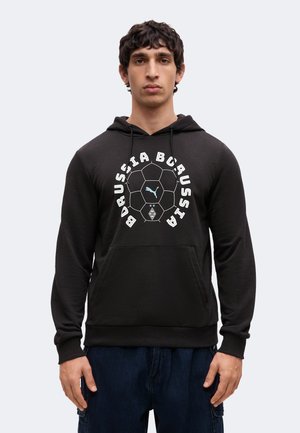 Jeune homme portant un sweat à capuche noir avec un motif ballon de football et le texte "Borussia Borussia", associé à un pantalon bleu foncé.