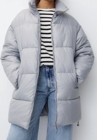 Chaqueta acolchada gris claro con cuello alto, cierre con cremallera y costuras en forma de diamante; llevada sobre una camiseta de rayas en blanco y negro.