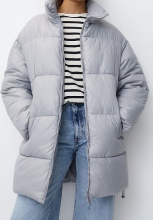 Lys grå pufferjakke med høj krave, lynlås foran og diamantformet syning; båret over en sort-hvid stribet skjorte.