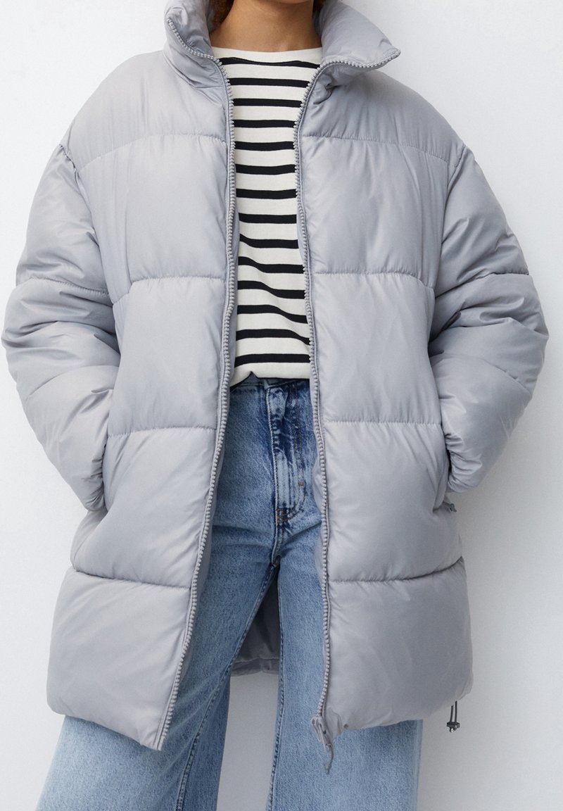 Chaqueta acolchada gris claro con cuello alto, cierre con cremallera y costuras en forma de diamante; llevada sobre una camiseta de rayas en blanco y negro.