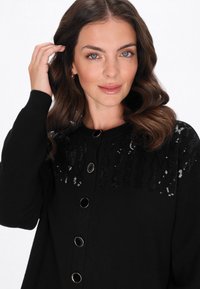 Cardigan nero con una parte superiore texturizzata e dettagli in paillettes, completo di bottoni tondi neri. Tessuto morbido e vestibilità comoda per il massimo del comfort.