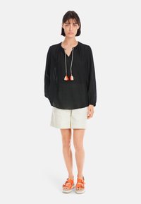 Schwarze Leinenbluse mit V-Ausschnitt und Kordelzug, akzentuiert durch bunte Quasten, kombiniert mit cremefarbenen Shorts und orangen Plateausandalen.