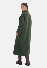 Abrigo largo verde con un diseño estructurado, cuello ancho y mangas sueltas. Combinado con botas marrones hasta la rodilla. Textura suave, sin cierres visibles.