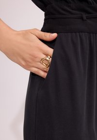 Czarne spodnie z paskiem i bocznymi kieszeniami. Na kieszeni spoczywa ręka, na której znajduje się złoty pierścionek o teksturowanej powierzchni. Gładki materiał o lekkim połysku.