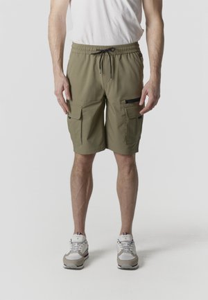 Olíva zöld, több zsebes cargo rövidnadrágot, fehér tornacipőt és fehér pólót viselő férfi, egyszerű háttér előtt állva.