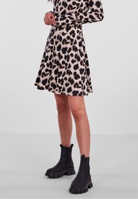 Kvinna klädd i en leopardmönstrad kjol och matchande topp, i kombination med svarta boots med kraftig sula, står framför en enfärgad bakgrund.