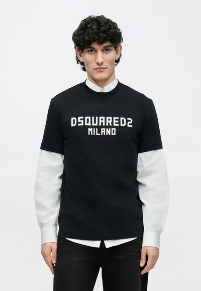 Jauns vīrietis ar cirtainiem matiem valkā melnu DSQUARED2 Milano T-kreklu virs baltas garām piedurknēm krekla uz vienkrāsaina balta fona.