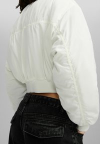 Krótkia biała kurtka puchowa o gładkim, błyszczącym wykończeniu i elastycznych mankietach. Widoczne w talii czarne, wysokowodne dżinsy.