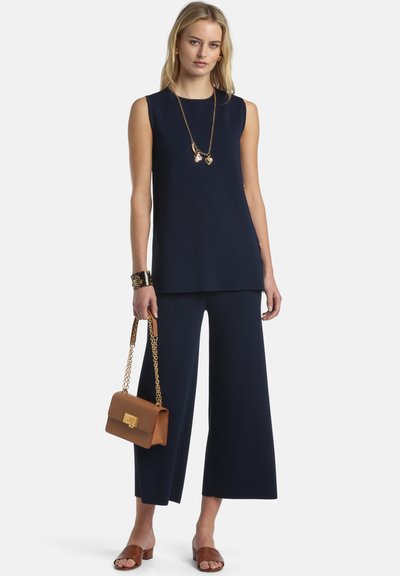 Femme blonde portant un haut bleu marine sans manches et un pantalon à jambes larges, tenant un sac à main beige avec une chaîne dorée, accessoirisée d'un collier et d'un bracelet en or.