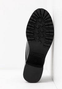 Semelle de chaussure noire avec un motif de bande de roulement profond incluant des formes croisées et des grips rectangulaires, inclinée contre un arrière-plan blanc en coin.
