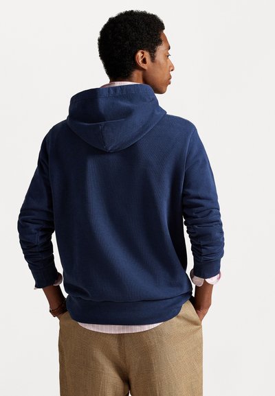 Temno modra kapuca majica iz soft materiala, z dolgimi rokavi in sproščenim krojem. Nošena z lahkotno karirasto srajco pod njo.