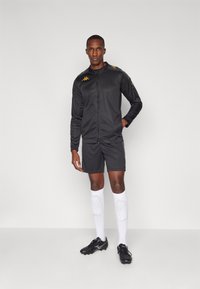 Homme en veste de sport noire et shorts, portant des chaussettes blanches jusqu'aux genoux avec protège-tibias, et des chaussures de sport noires, se tenant contre un fond clair.