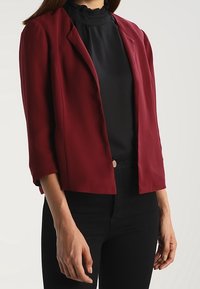 Cropped burgundy blazer with structured shoulders, satin finish, and three-quarter sleeves worn over a black ruffled neck top and dark jeans.