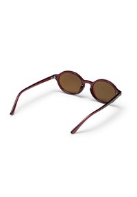 Στρογγυλά γυαλιά ηλίου με σκούρους καφέ φακούς και διαφανή σκούρα κόκκινα πλαίσια που ακουμπούν ανοιχτά σε λευκή επιφάνεια.