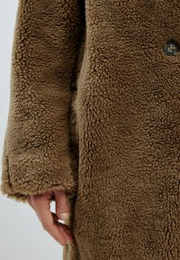 Brauner Kunstpelzmantel mit einer strukturierten, flauschigen Außenseite, ausgestattet mit einem Knopfdetail und weiten Ärmeln. Eine Hand ruht an der Seite des Mantels.