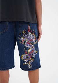 Denim shorts featuring a large embroidered dragon design in purple, red, and yellow on the right leg. Dark blue color with visible stitching.