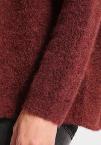Burgunderfärgad stickad tröja med en mjuk, fluffig textur och breda, ribbade manschetter. Tyget ser varmt och mysigt ut, perfekt för kyligare väder.