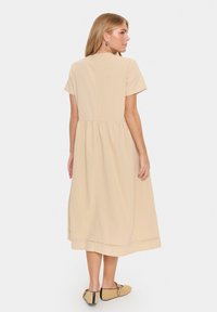 Rochie bej, cu mâneci scurte, cu un corset ajustat și talie strânsă, care coboară până la jumătatea gambei, având un tiv simplu. A fost purtată cu pantofi slip-on de culoare bej.