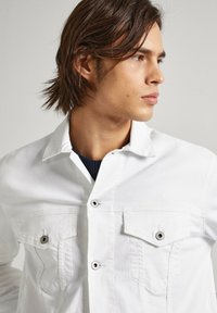 Witte denim jack met twee borstzakken, zilveren knoopaccenten en een klassieke kraag. De stof heeft een subtiele textuur.