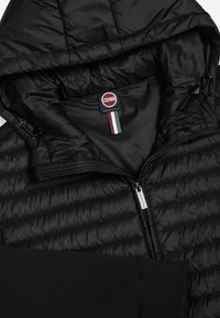 Czarna pikowana kurtka z kapturem, z przodu zamek błyskawiczny, ściągacze na mankietach i widoczna metka Colmar Originals wewnątrz kołnierza.