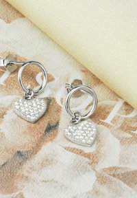 Silverfärgade hoopsmedhjälpörhängen med hjärtformade mönster, smyckade med klara kristaller, uppvisade mot en mönstrad tygbakgrund.