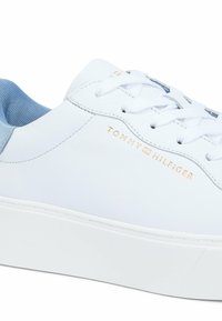 Sneaker in pelle bianca con dettagli in suede azzurro chiaro, suola piatta in gomma bianca e logo dorato "Tommy Hilfiger" sul lato.