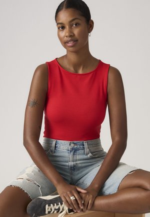 Žena u crvenom topu bez rukava i poderanim traper kratkim hlačama, sjedi prekriženih nogu u crnim tenisicama na drvenoj površini.