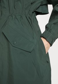 Zelená nepromokavá bunda s elastickým pasem, kapsou na patentky a nařasenými manžetami. Hladká textura s minimalistickým designem.