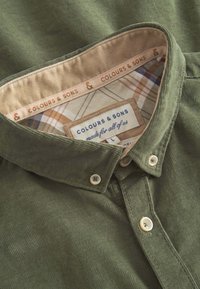 Camicia di velluto a coste verde oliva con colletto button-down, caratterizzata da una fodera interna a quadri e etichetta "Colours & Sons" sul colletto.