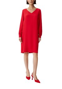 Vestido rojo de manga larga con escote en V, fabricado en una tela suave. Presenta puños fruncidos y un dobladillo a la altura de la rodilla. Combinado con zapatos de tacón rojos.