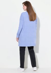 Kvinna med rakt, långt brunt hår iförd en ljusblå oversized tröja, svarta byxor och vita sneakers, stående mot en vit vägg.