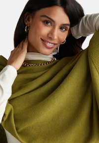 Poncho a maglia verde oliva con una morbida texture, caratterizzato da tessuto drappeggiato e un collo alto. Accentuato da una collana con catena dorata.