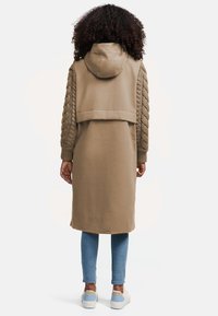 Jachetă de culoare brown cu glugă, având mâneci texturate cu model de tricot. Haina are o lungime medie, fiind asortată cu blugi albaștri și încălțăminte deschisă.
